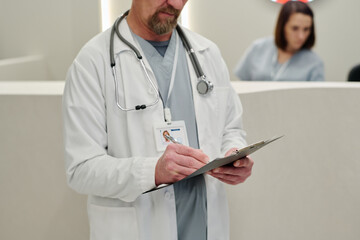 Experienced clinician with medical document and stethoscope making notes against reception counter with young female nurse