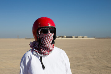 Man wearing keffiyeh and a helmet wrapped around his face stands against the backdrop of desert...