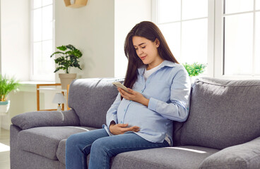 Young pregnant woman sitting on the sofa at home, holding her mobile phone, using a pregnancy week...