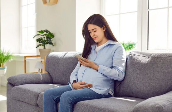 Young pregnant woman sitting on the sofa at home, holding her mobile phone, using a pregnancy week by week tracker app, looking at an ultrasound image of her baby or photos of children on social media