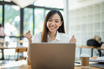 A delighted Asian businesswoman raises her fists in victory while looking at her laptop in a well-lit office space..