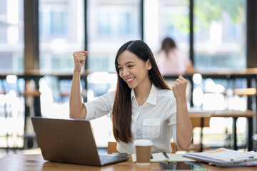 A delighted Asian businesswoman raises her fists in victory while looking at her laptop in a...