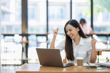 A delighted Asian businesswoman raises her fists in victory while looking at her laptop in a well-lit office space..