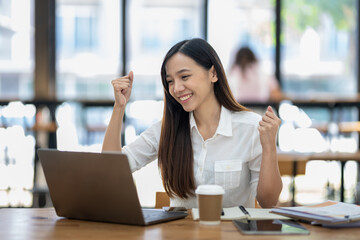A delighted Asian businesswoman raises her fists in victory while looking at her laptop in a well-lit office space..