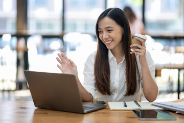 A friendly Asian businesswoman waves hello during a video call on her laptop, holding a coffee cup in a bright office setting..