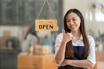 Asian female cafe owner with a welcoming smile presents the open sign at her shop, signaling readiness for customers..