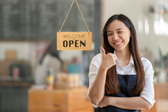 Asian female cafe owner with a welcoming smile presents the open sign at her shop, signaling readiness for customers..