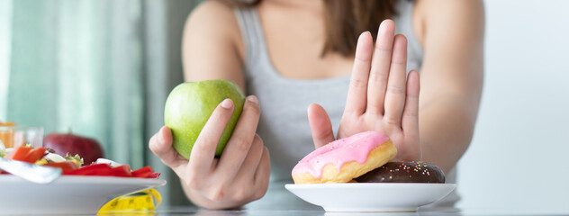Woman on dieting for good health concept. Close up female using hand push out her favourite donut and choose green apple and vegetables for good health. - obrazy, fototapety, plakaty