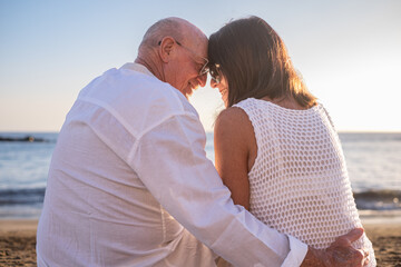 Rear view of bonding senior couple hugging while sitting on the beach face the sunset enjoying...