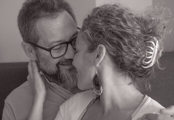 Black and white portrait of lovely mature couple sitting on home sofa hugging each other tenderly, exchanging cuddles and kiss, concept of love, protection and care