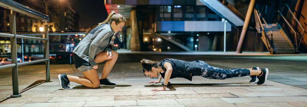 Young woman doing push ups during hard training by supervision of her female personal trainer on the empty city at night
