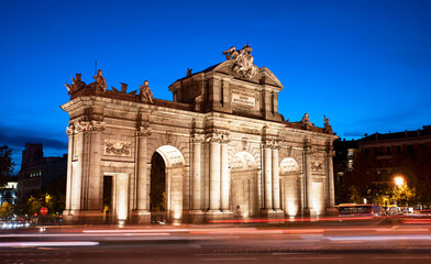 Obraz premium Puerta de Alcalá by the entrance of El Retiro park at blue hour in Madrid. Puerta Alcala evening view with car light trails as the night approaches. Downtown vibe on the Spanish capital busy streets