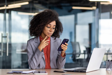 Focused woman with curly hair using smartphone while working in a contemporary office setting, showcasing multitasking and efficiency.