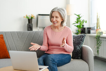 Attractive smiling senior woman using laptop computer having video call, communication online, explaining something sitting on comfortable sofa at home. Gray haired businesswoman working from home