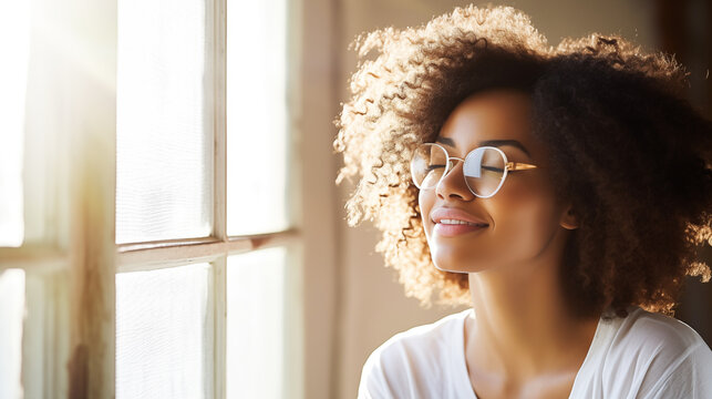 Woman in white t-shirt and glasses, standing by the window with gentle sunlight casting a warm glow, eyes closed and a subtle smile, embodying serene joy