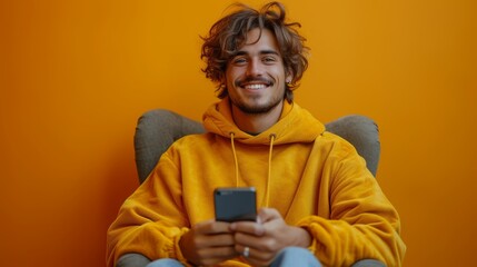 A smiling young man using his smartphone sitting on his chair isolated on an orange studio background. He is chatting online and browsing social media.