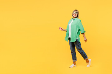 Fototapeta premium Full body elderly side profile view blonde woman 50s years old wear green shirt glasses casual clothes walking going look camera isolated on plain yellow background studio portrait. Lifestyle concept.