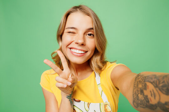 Close up young housewife housekeeper chef cook baker woman she wears apron yellow t-shirt do selfie shot on mobile cell phone show v-sign wink isolated on plain green background. Cooking food concept.