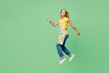 Full body side view smiling cheerful young housewife housekeeper chef cook baker woman wear apron yellow t-shirt jump high look camera isolated on plain green background studio. Cooking food concept.