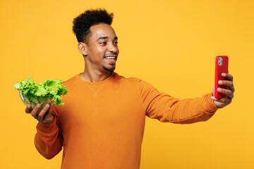Young man wear casual clothes hold fresh greens lettuce leaves in bowl do mobile cell phone selfie shot isolated on plain yellow background Proper nutrition healthy fast food unhealthy choice concept
