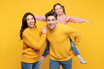 Young parents mom dad with child kid girl 7-8 years old wear pink knitted sweater casual clothes giving piggyback ride to joyful, sit on back isolated on plain yellow background. Family day concept.