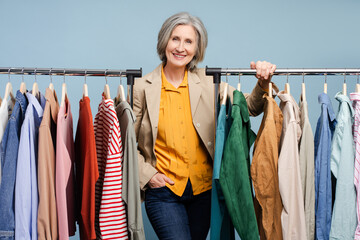 Smiling confident senior woman, stylist, fashion designer standing near clothes rail with a lot stylish clothes. Portrait successful tailor in atelier. Shopping, store, small business concept