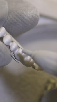 Vertical video. Dental technician polishing artificial ceramic teeth using sandblaster in the dental lab. Professional wearing gloves while holding fake teeth in hand and polishing it with sand