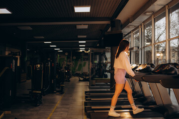 Obraz na płótnie Canvas Woman running on treadmill at a panoramic window and listening to music via headphone at gym. Concept of healthy lifestyle.
