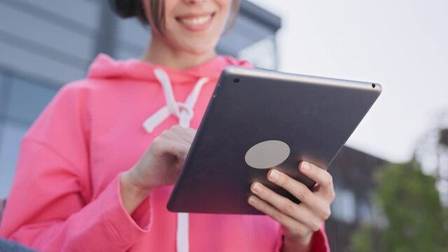 Close up of unknown woman with short haircut in pink sweatshirt typing on digital tablet on blurred city background. Female gadget user chatting online in messenger with friends while having a walk.