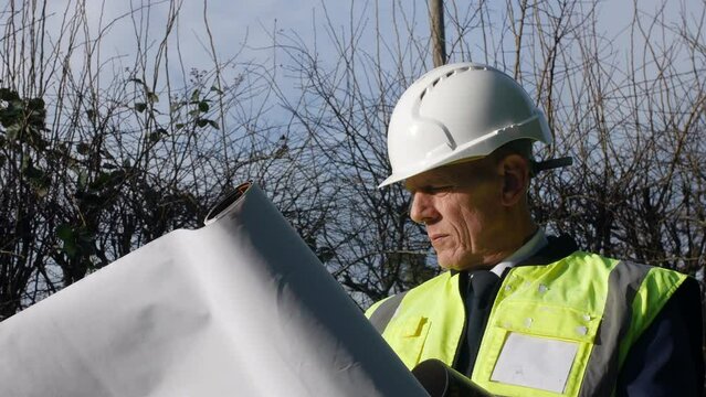 Close up portrait of an mature senior architect building inspector inspecting a construction site with architectural plans and hard hat