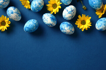 Frame border of blue and white Easter eggs on dark blue background with yellow flowers. Flat lay, top view, copy space.