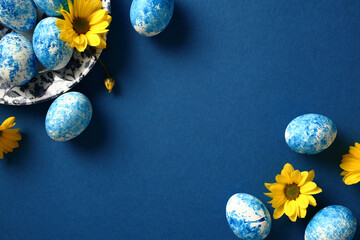 Blue and white handmade Easter eggs with yellow flowers on dark blue background. Flat lay, top view.