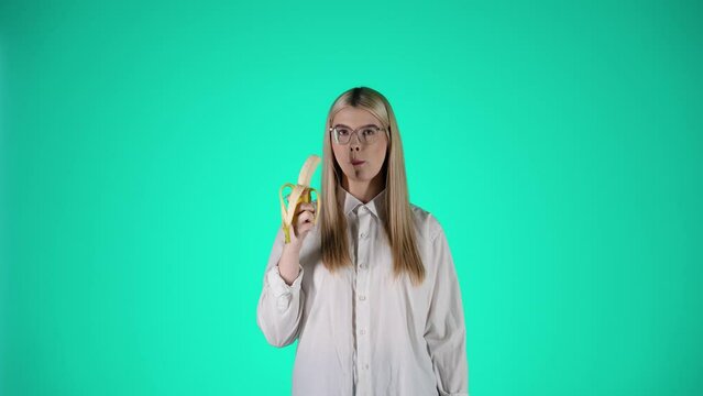Młoda blond kobieta w białej koszuli i okularach jedząca banana i uśmiechająca się do kamery &mdash; centralne ujęcie na zielonym tle, naturalność i smak w slow motion 4K
