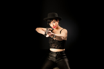 Fototapeta na wymiar Young mentally unstable and paranoid woman with a gun in a black hat on a black background