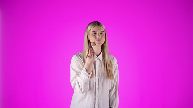 Blondynka w okularach i białej koszuli na r&oacute;żowym tle delikatnie nakłada puder na twarz i nos. Spokojne, kobiece ujęcie ukazujące moment codziennej pielęgnacji i dbania o urodę