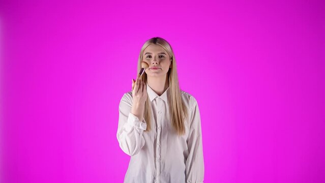 Młoda kobieta w białej koszuli z blond rozpuszczonymi włosami pozująca z uśmiechem i pudrująca twarz pędzlem do makijażu &mdash; r&oacute;żowe tło, beauty, styl i kobiecość 4K