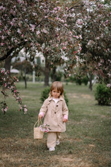 A little girl against the background of an apple tree blooming with red flowers. A girl holds a basket of apple flowers in her hands. The child is dressed in a peach raincoat and white sneakers