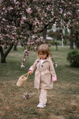 A little girl, a child in a beige raincoat and trousers walks along a green lawn in a garden of blooming apple trees. Spring