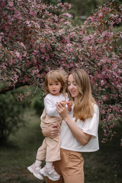 Beautiful mother and daughter against the background of a blooming apple tree. Mom holds her little daughter in her arms. Stylish clothes in neutral colors. Spring day in the park