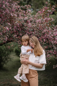 Beautiful mother and daughter against the background of a blooming apple tree. Mom holds her little daughter in her arms. Stylish clothes in neutral colors. Spring day in the park