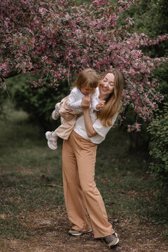 Beautiful mother and daughter against the background of a blooming apple tree. Mom holds her little daughter in her arms. Stylish clothes in neutral colors. Mom and daughter having fun