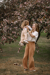Beautiful mother and daughter against the background of a blooming apple tree. Mom raises her little daughter in her arms. Stylish clothes in neutral colors. Mom and daughter having fun