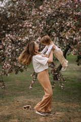 Beautiful mother and daughter against the background of a blooming apple tree. Mom raises her little daughter in her arms. Stylish clothes in neutral colors. Mom and daughter having fun