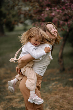 Beautiful mother and daughter against the background of a blooming apple tree. Mom rides her little daughter on her back. Stylish clothes in neutral colors. Mom and daughter having fun