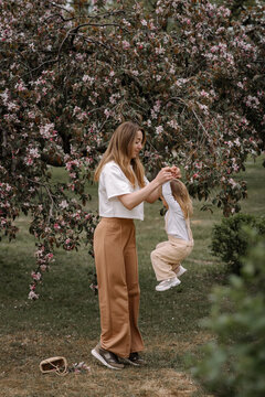 Beautiful mother and daughter against the background of a blooming apple tree. Mom holds her little daughter's hands. Stylish clothes in neutral colors. Mom and daughter having fun