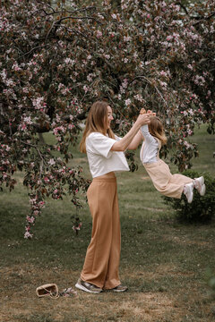 Beautiful Mother And Daughter Against The Background Of A Blooming Apple Tree. Mom Holds Her Little Daughter's Hands. Stylish Clothes In Neutral Colors. Mom And Daughter Having Fun