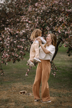 Beautiful Mother And Daughter Against The Background Of A Blooming Apple Tree. Mom Raises Her Little Daughter In Her Arms. Stylish Clothes In Neutral Colors. Mom And Daughter Having Fun