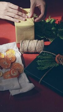Vertical. A hand wrapping a gift box with recycled paper amidst twine, pine leaves and dried orange slices for decoration, creative and sustainable idea