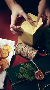 Vertical overhead view of hands wrapping a gift box with recycled paper surrounded by twine, dried slices of citrus and pines to be used as decoration showing creativity and sustainability