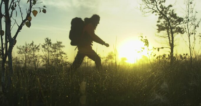 Man with dreadlocks and a backpack walking in a raised bog during foggy sunrise. Dew grass in foreground. Slow motion.